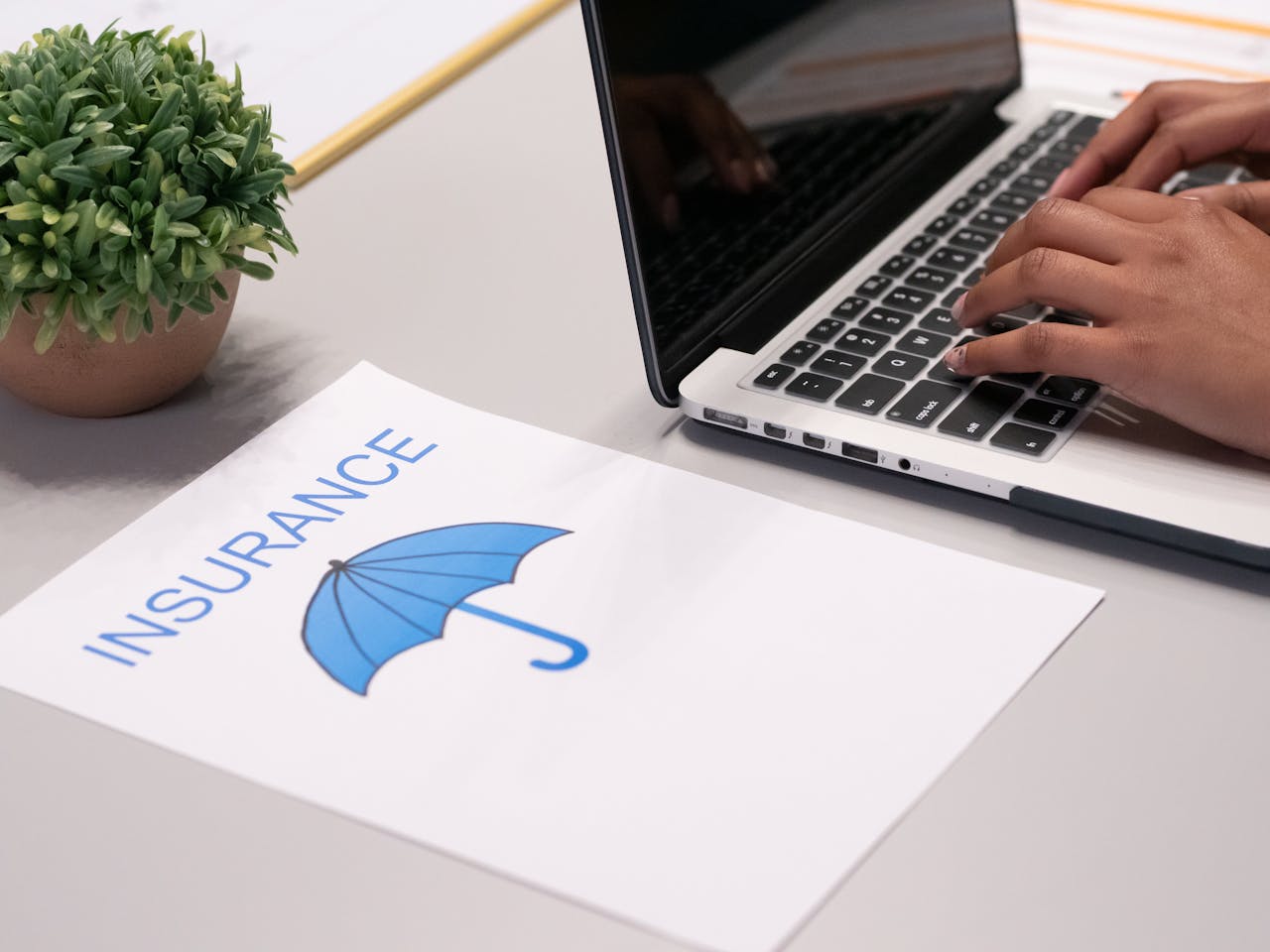 A person typing on a laptop next to a paper that says insurance above an umbrella.