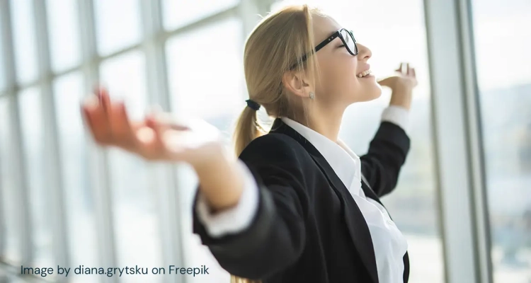 Smiling business woman stands in front of windows with closed eyes and outstretched arms