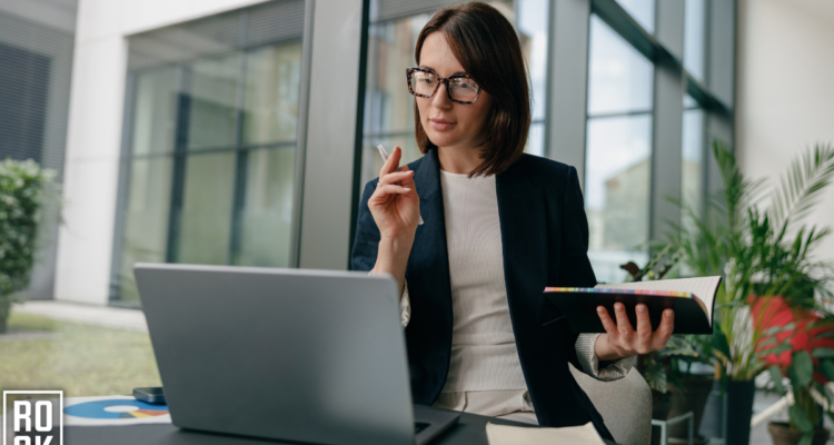 women working on a laptop while holding a notebook in her hand