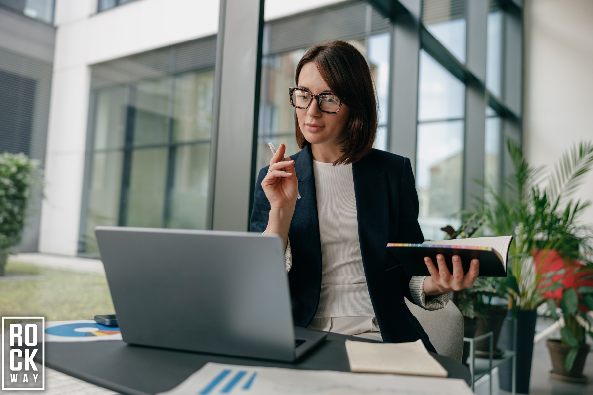 women working on a laptop while holding a notebook in her hand